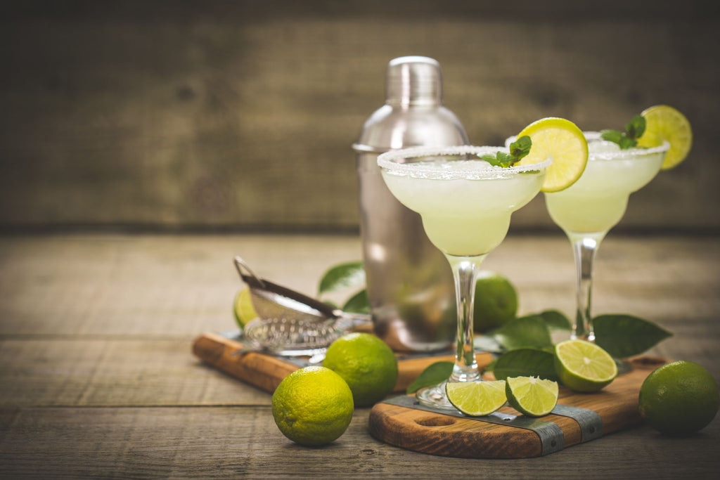 Two margaritas on a cutting board next to limes a strainer, and a cocktail shaker 