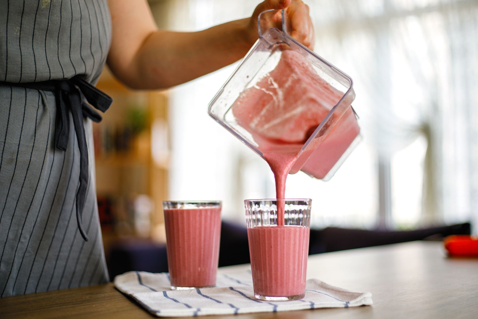A person pours a blended pink smoothie into glass cups.