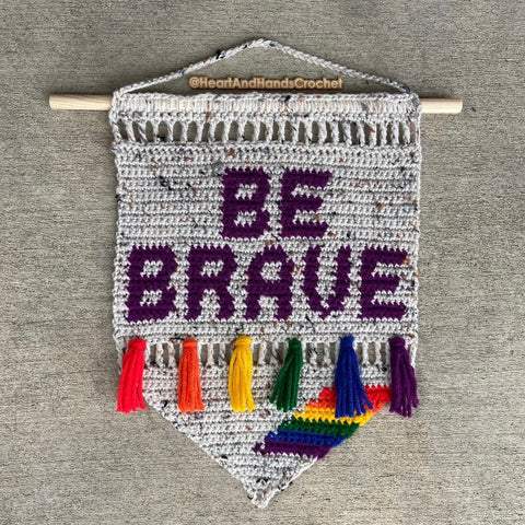 Crochet banner hanging from a dowel rod with the words BE BRAVE in the design, plus 6 tassels in rainbow colors and 6 small stripes at the bottom in the same rainbow colors