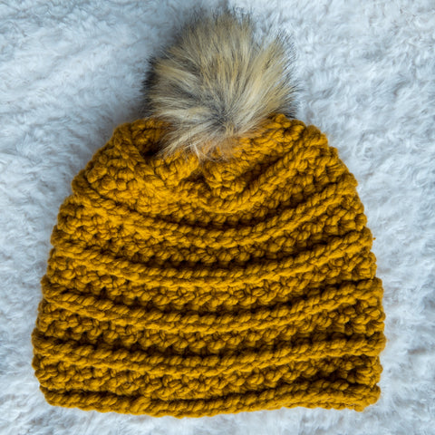 Mustard-colored crochet beanie with ridged texture and a faux fur pom