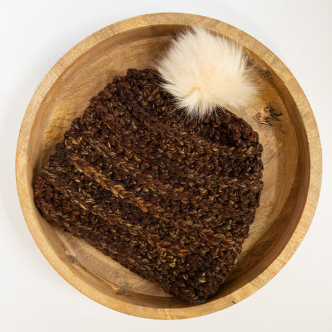 A deep brown speckled crocheted beanie with an ivory pom resting in a brown wooden tray on a white background