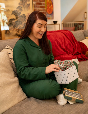 A woman in cozy, breathable pajamas sits on a couch, smiling as she opens a gift box.