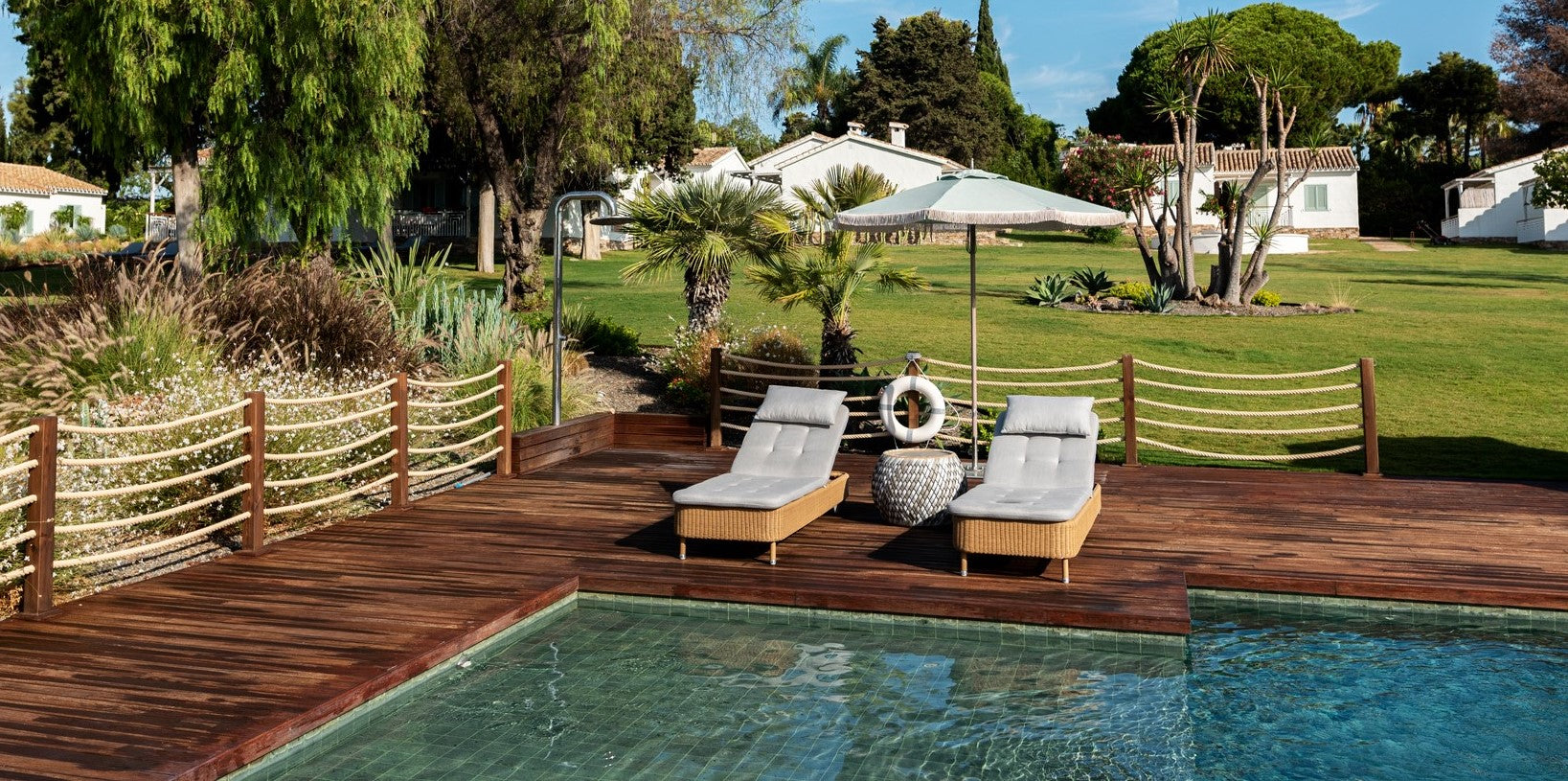 two light brown sunbeds by the poolside under a parasol with round side table surrounded by nature