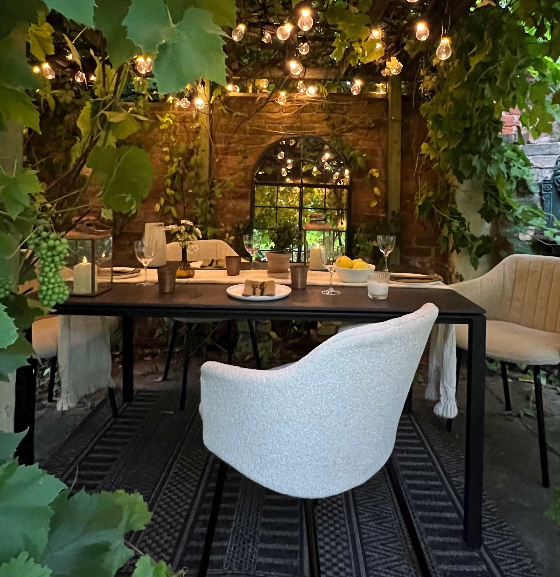 Back of a white bouclé garden chair with a dining setup under a lush green pergola in the summer times