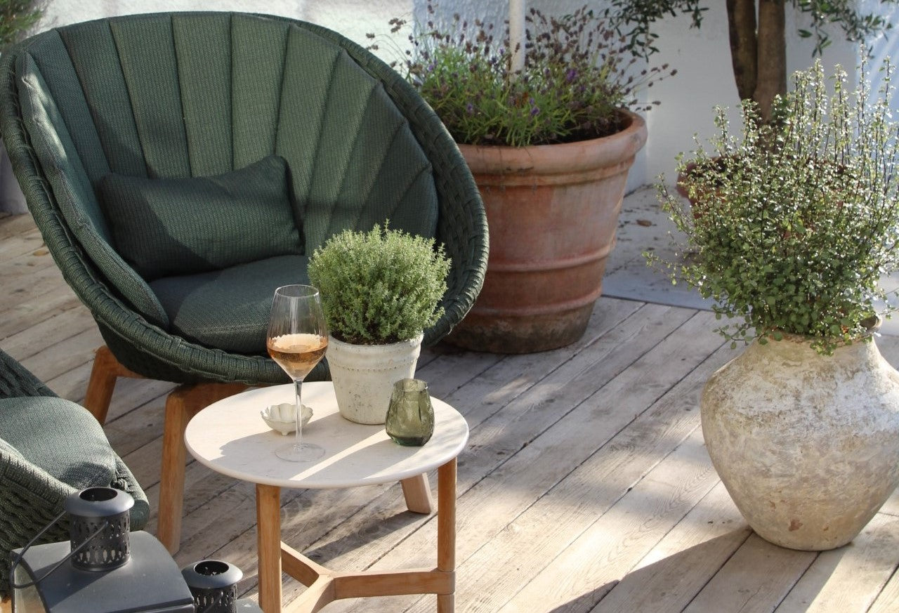 Front of Cane-line Peacock lounge chair in dark green colour with a small coffee table white white travertine ceramic table top and teak base. Glass of rose, small plant and candlelight