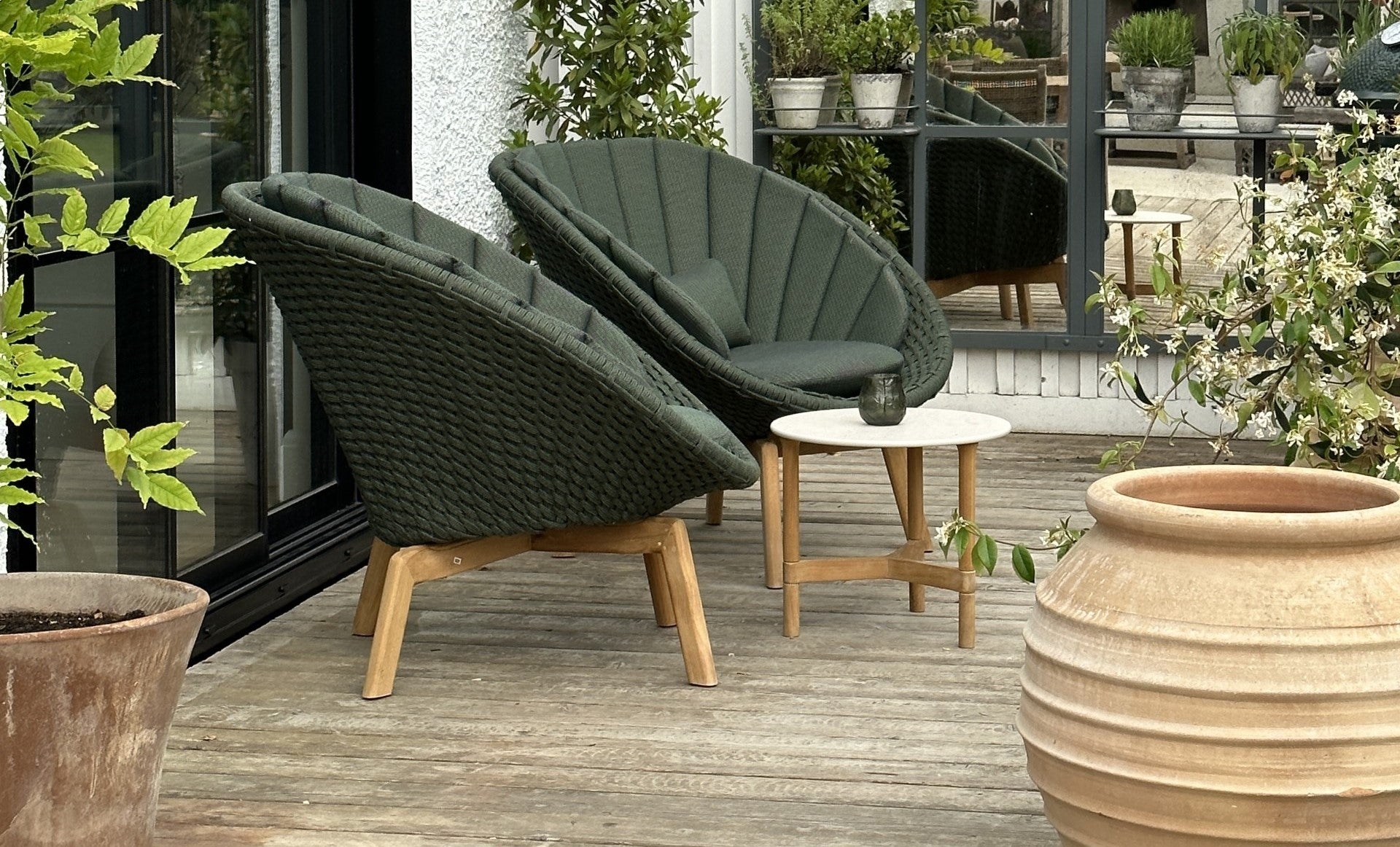 Two dark green Cane-line Peacock lounge chairs with a shell-shaped form, teak base, white travertine ceramic side table with teak base, green plants allround, ceramic pots