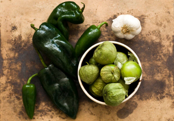 top view shot of 5  jalapeños beside 1 whole garlic, and a bowl of green tomatillos on a wooden surface.