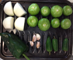 1 big white onion divided into 4 slices, 8 unwrapped tomatillos, 1 poblano chili, 4 cloves of garlic, and 3 jalapeño on a black pan