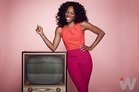 Yvonne orji in a coral top and dark pink trousers posed next to an old-style television.