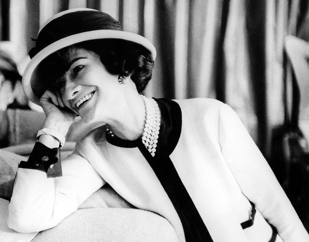 coco chanel, wearing a hat smiling, whilst leaning on to a table.