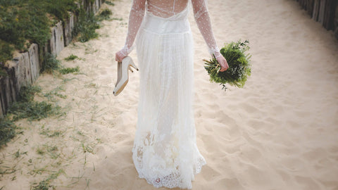 bride walking along the beach carrying her shoes 