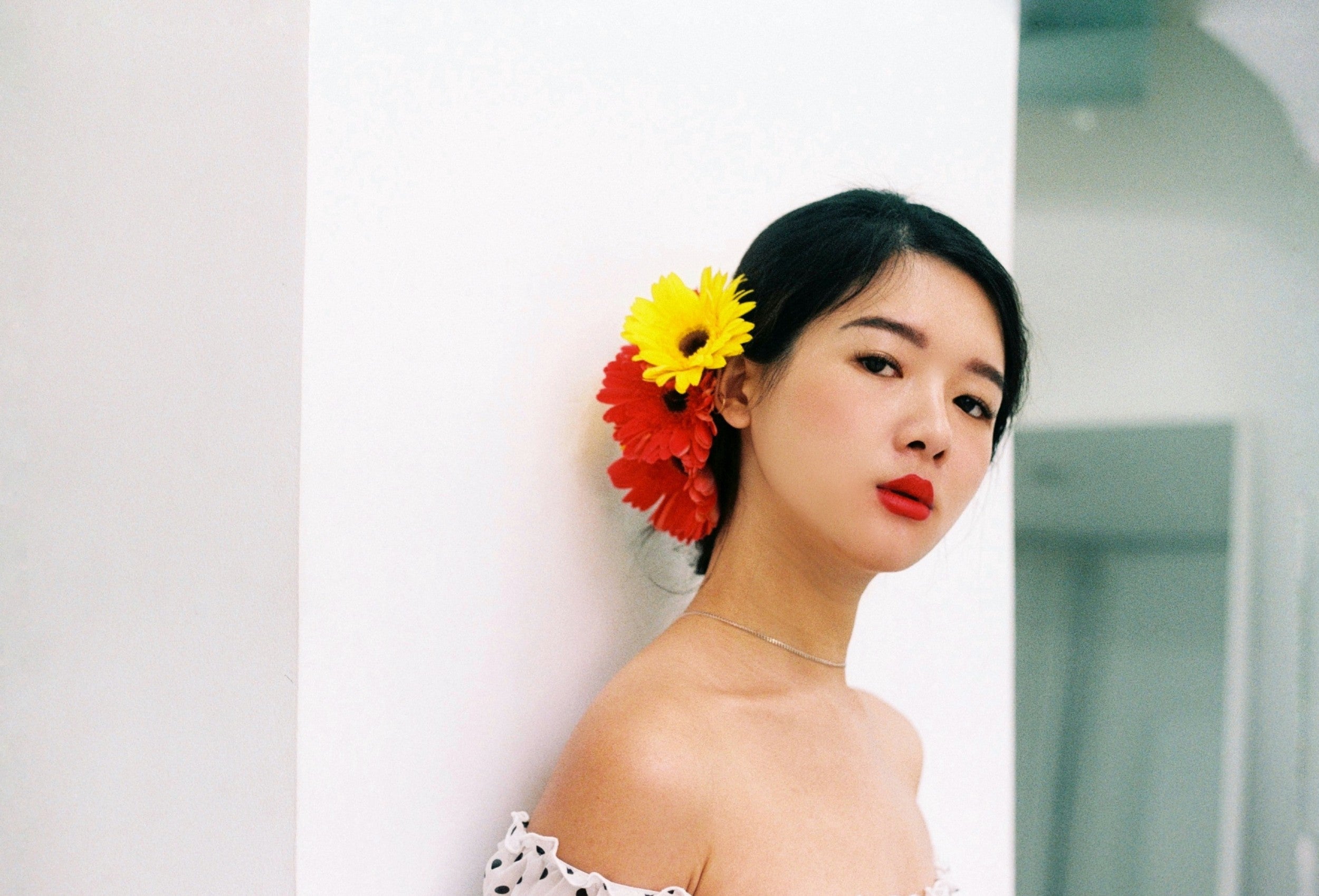 a woman pictured leaning against a white wall, wearing a low cut polka dot dress with bright yellow and red flowers in her tied back hair.