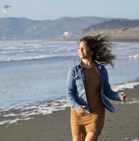 a woman in a brown short dress and blue jean open shirt is running on a beach with the water behind her. her hair is flying in the wind