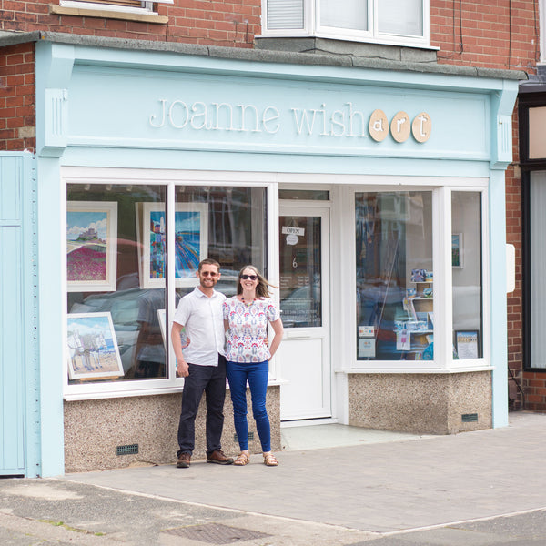 Small business owners Ross and Joanne Wishart stand outside their  pale blue gallery shop front. The shop windows are displaying some colourful paintings.