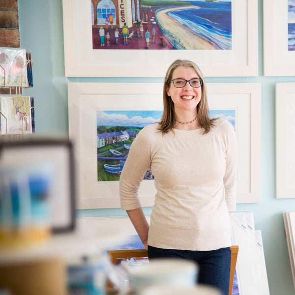 Lady stands in her art gallery with paintings on the wall behind her. 
