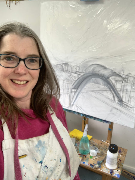 Artist Joanne Wishart is standing in front of a canvas on an easel. There is the beginnings of a painting of the Tyne Bridge. 