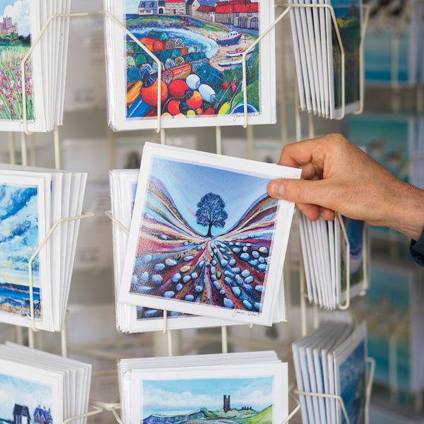 A hand is pulling a greetings card out of a card rack. THe card features and image of a tree. 