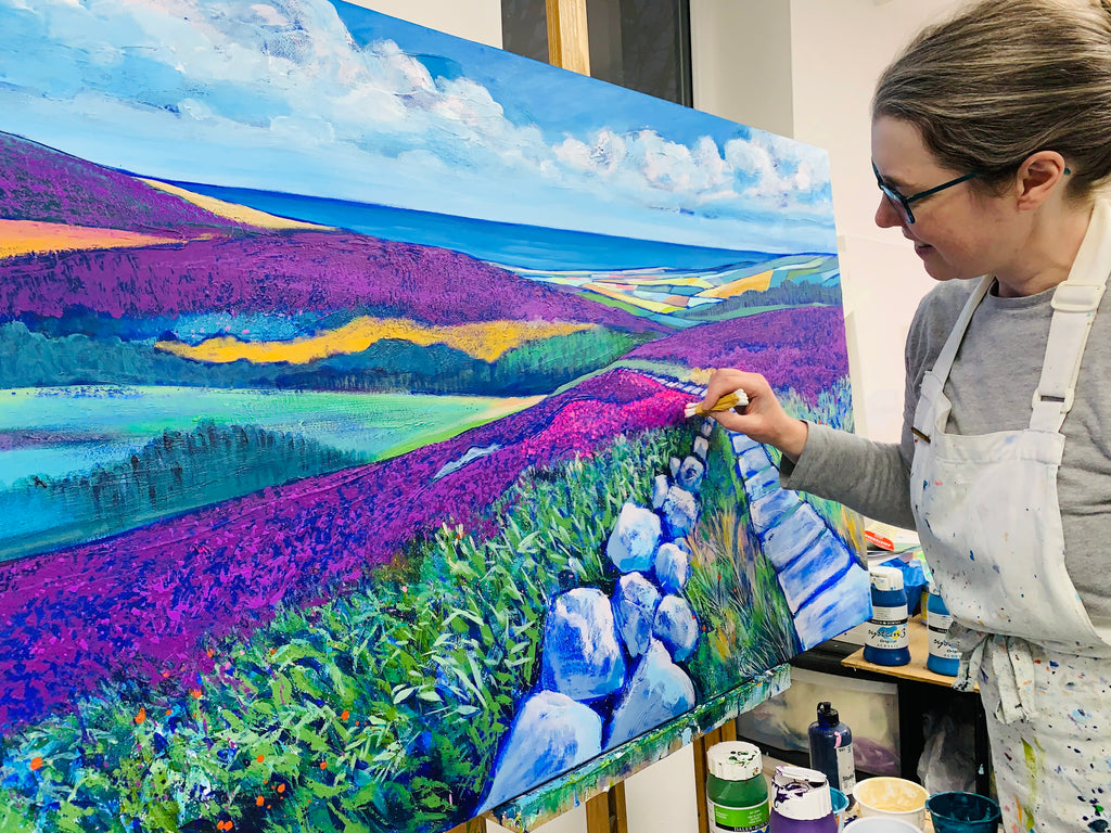 Artist stands painting at her easel. The painting is of a purple landscape scene looking out towards the sea. 