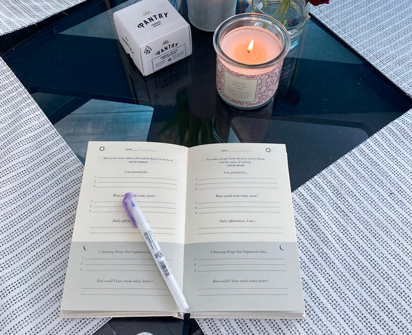 open gratitude journal showing a few writing prompts. set on a table with a candle and pen
