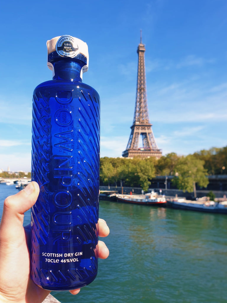 A hand holds up a blue Downpour bottle in front of the Eiffel Tower. The sky is blue and bright and the Seine shines turquoise below.