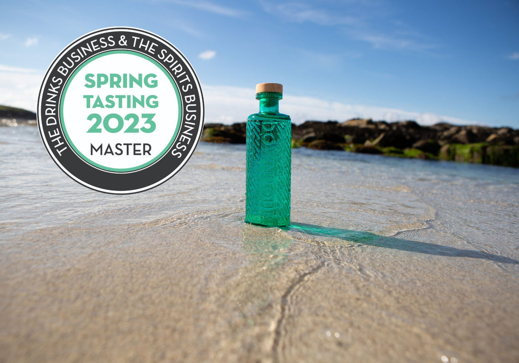 A turquoise bottle of Downpour gin sits on a pale sand shore, with water lapping shallowly around it. The sky is blue and there are seaweed covered rocks in the background. There is a white medal with turquoise writing reading 'spring tasting 2023 MASTER' on top of the image 