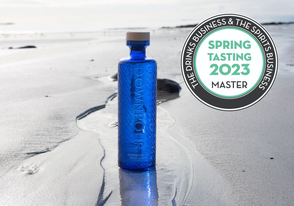 a deep blue bottle of Downpour Gin sits on silvery sand with rivulets of water running over it. A white and black medal with turquoise writing, reading 'Spring Tasting 2023 MASTER' sits on top of the image