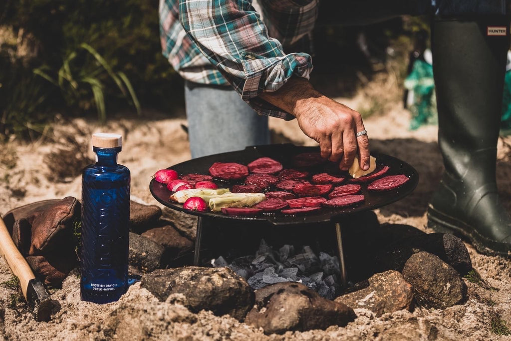 Beetroot cooking on a plancha over an open fire, on the beach. A bottle of Downpour gin sits next to it and the chef tends the beetroot