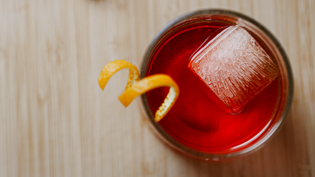 a glass filled with negroni, shot from above. The glass sits on a pale wooden surface, a square ice cube floats in the liquid, and a twist of orange peel is balance on the edge of the glass. 