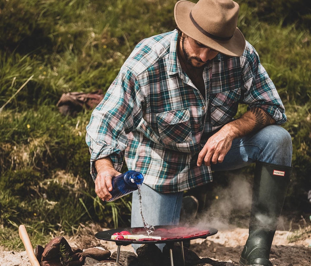 A man in a checked shirt and hat, leans over a griddle over an open fire. He pours gin from a blue Downpour bottle onto the sliced beetroot cooking on the griddle