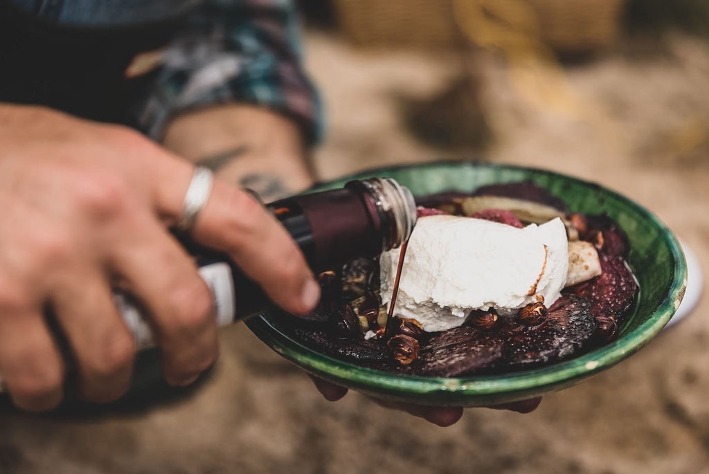 A pair of white hands holds a green plate, filled with grilled vegetables and ricotta cheese. One hand pours a dark red liquid from a bottle over the vegetables. 