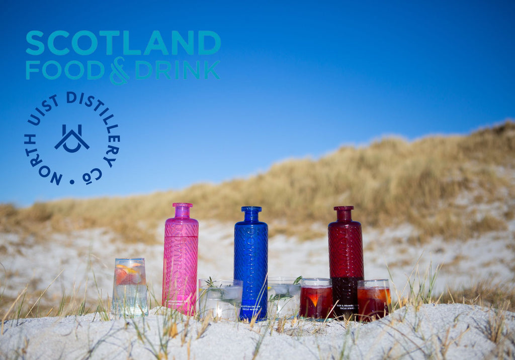 North Uist Distillery Downpour Gins, Pink Grapefruit, Scottish Dry, Sloe and Bramble, sitting on a white sand beach with a blue sky above them. 