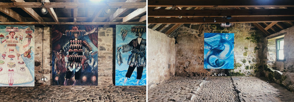 Two images side by side of large scale oil paintings in a cobbled stable space with wooden ceiling beams. The paintings are in shades of bright, vibrant blues and depict fantastical figures. 
