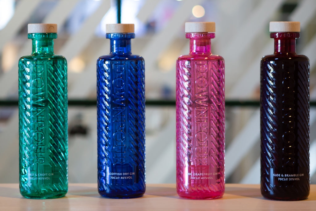 Four jewel bright bottles in different colours (turquoise, cobalt, barbie pink, and deep blood red) sit on a plywood bar. The bottles have straight sides and are embossed with rain pattern. They have wooden stoppers and the word Downpour stands out on the front.