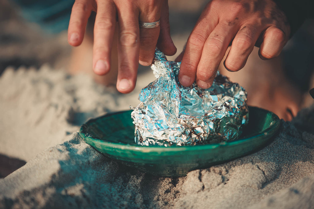 a close up of a pair of white hands, unwrapping a foil package sitting on a green plate. The plate sits on the sand. 