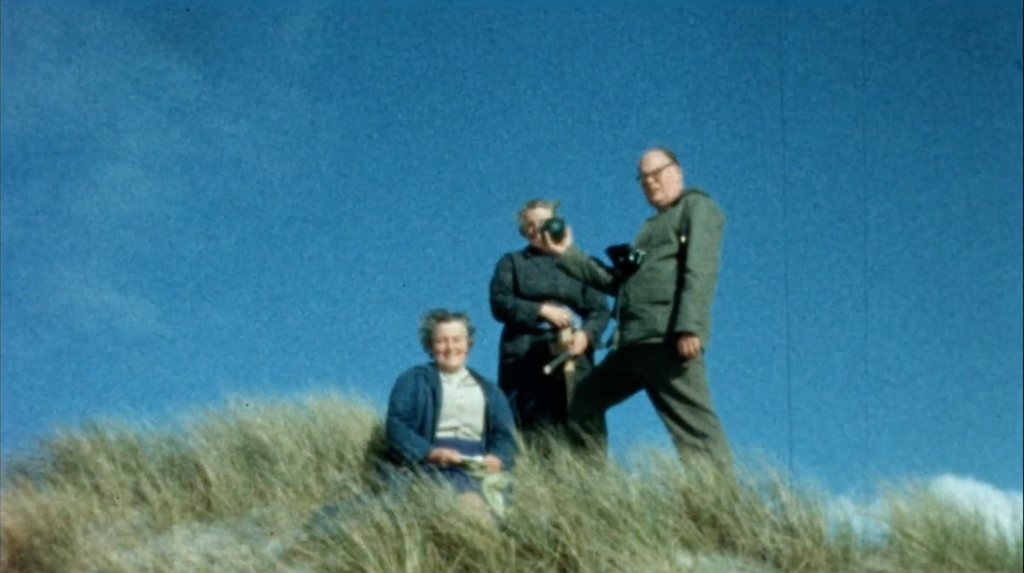 still from a vintage film reel, three people standing in a sand dune, with long grass in front of them and blue skies behind them 
