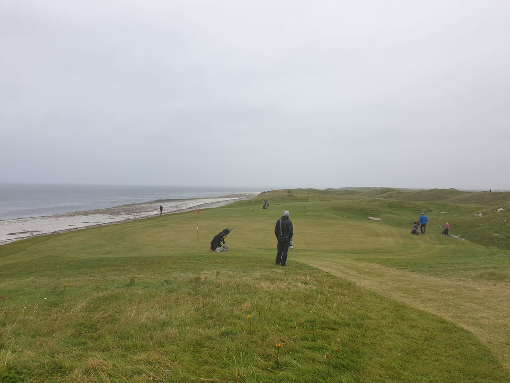 golfers stand on the green in the rain, the course sitting alongside a white sand beach 