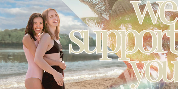 Two queer people hugging wearing trans swimwear with text that says, "we support you."
