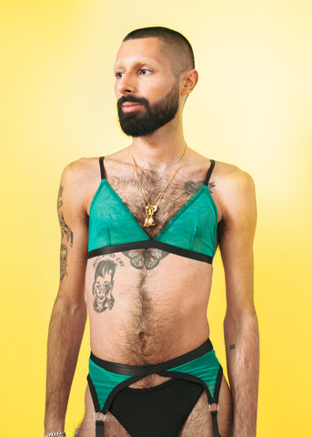 Masc queer person wearing fem green mesh lingerie against a yellow background
