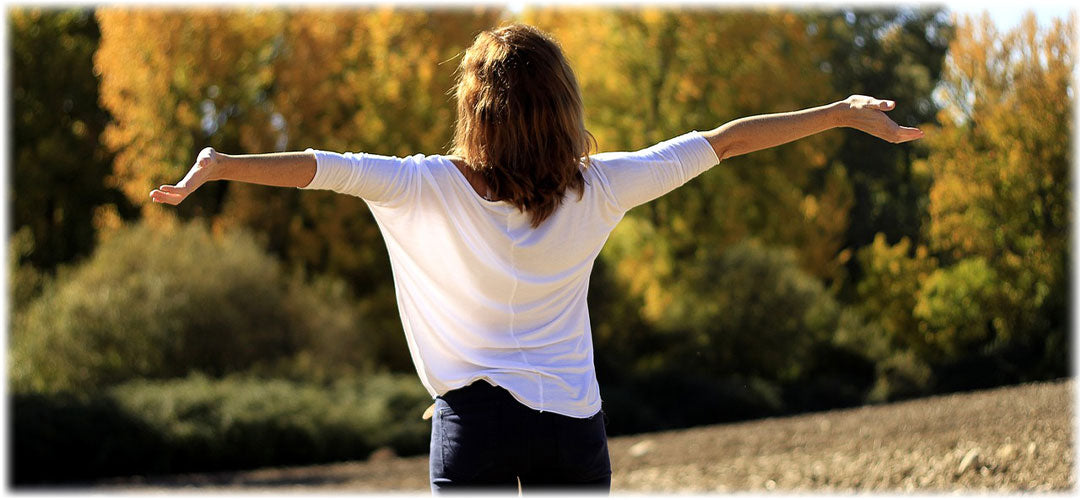Woman in a white shirt standing outdoors with her arms outstretched, surrounded by nature. The scene conveys a sense of freedom and well-being