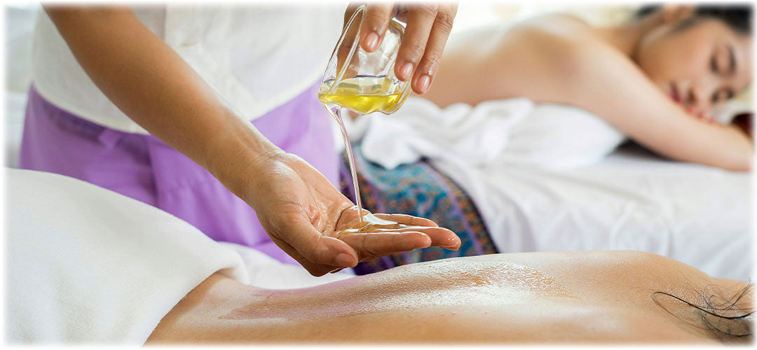 Massage therapist pouring essential oil into their hand before applying it to a client's back. Another client relaxes in the background. The scene exudes relaxation and rejuvenation