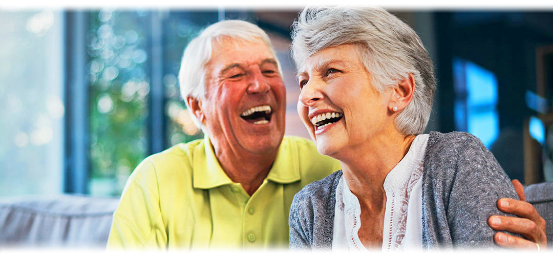 A group of older adults smiling and laughing while sitting together outdoors. They are wearing casual clothing and enjoying each other's company in a park or garden setting.