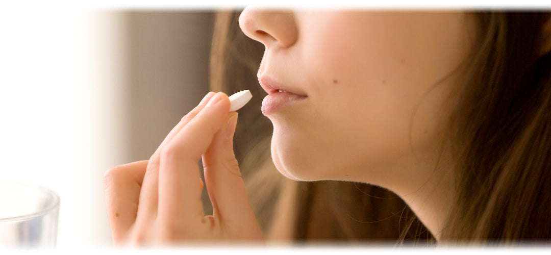 A person holding a small pill between their thumb and index finger, with a glass of water nearby. The focus is on the pill, suggesting the concept of taking medication or supplements.