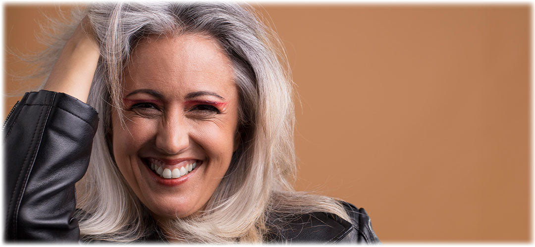 Smiling middle-aged woman with gray hair and vibrant eye makeup wearing a black leather jacket. Her joyful expression and hand in her hair convey confidence and positivity against a warm, neutral background.