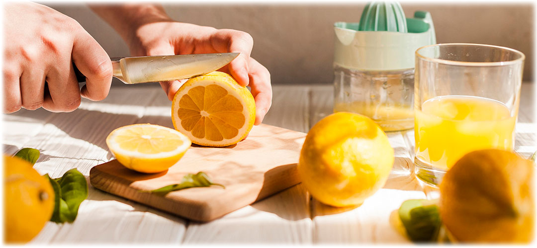 Person slicing a lemon on a wooden cutting board next to freshly squeezed lemon juice in a glass and a manual citrus juicer. Bright and sunny kitchen setting, highlighting the freshness and health benefits of citrus fruits.