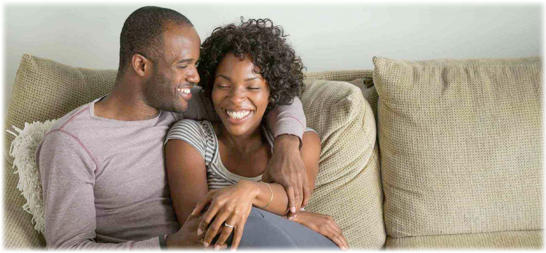 Smiling couple sitting closely on a beige sofa. The man has his arm around the woman, both looking at each other affectionately, conveying love and happiness.