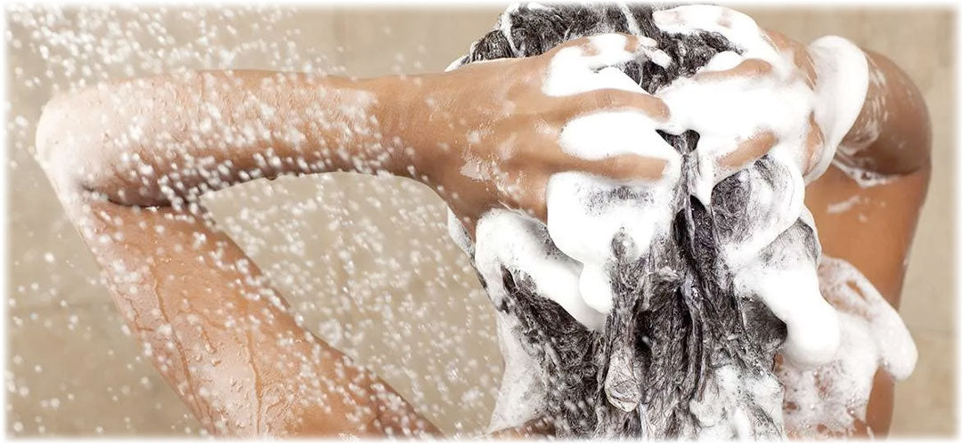 Person washing their hair in the shower, lathering up shampoo and surrounded by water droplets. The scene emphasizes cleanliness and hair care.