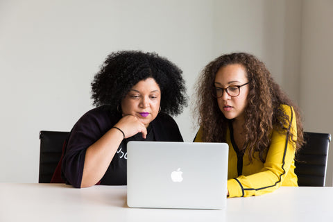 two women of color sitting at a table, collaborating together, looking at a apple macbook laptop screen