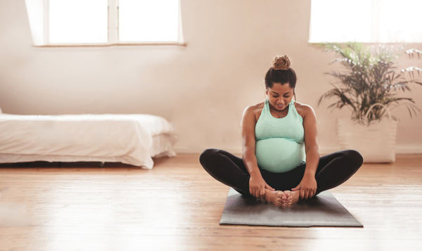 A pregnant woman doing yoga and stretching on the floor of an empty bedroom with a bed and windows