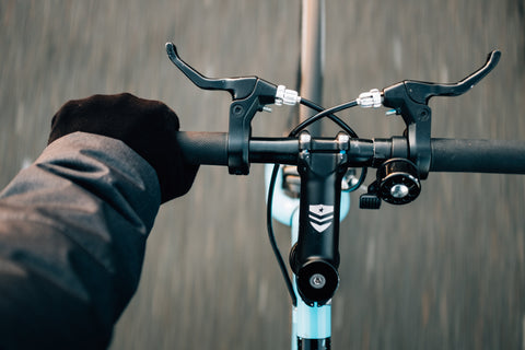 A close-up of a cyclist's hand wearing black gloves and operating the brake lever on a wet handlebar.