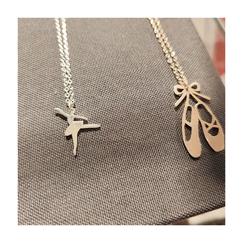 Two silver necklaces sitting side by side, one with a small ballerina charm and one with a pair of pointe shoes.